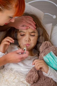 A little girl with her favorite teddy bear on whom she was wearing a gauze bandage is sick in bed and her mother measures her temperature with an electronic thermometer.
