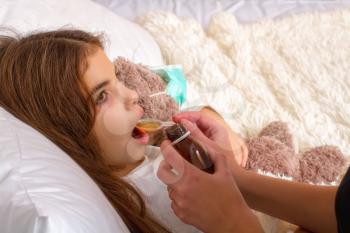 A little girl with a favorite teddy bear on whom they put on a gauze bandage is lying sick in bed and her mother is drinking her medicine.