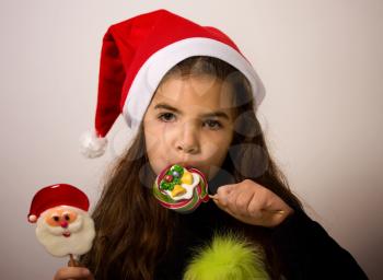 A little girl in a Santa Claus hat licks bright New Year's candies
