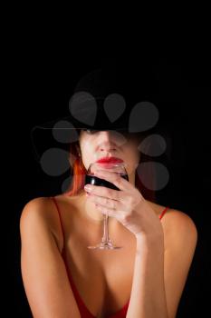 Sensual girl in a bright red dress with red hair drinks wine from a glass on a dark background
