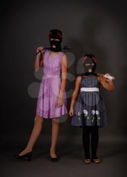 Two teenage hooligans in bright elegant dresses, but in gangster balaclava hats with heavy truncheons pose on a dark background