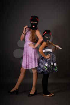Two teenage hooligans in bright elegant dresses, but in gangster balaclava hats with heavy truncheons pose on a dark background