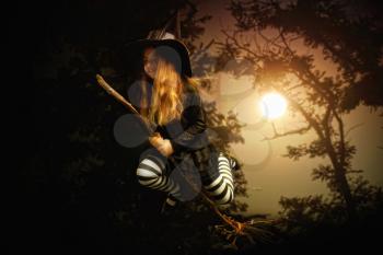 a little girl in traditional costume of a medieval witch and a pointed hat is flying on a broom through a dark forest