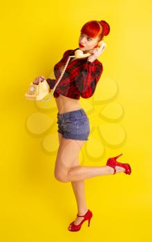 Pretty girl in knotted plaid shirt and shorts with retro phone talking to someone on a bright yellow background