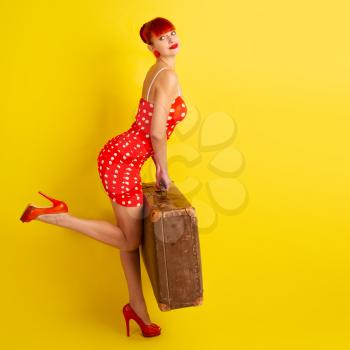 Pin up image Slender sexy girl in red dress in polka dot that holds in her hands a huge old suitcase on a bright yellow background.
