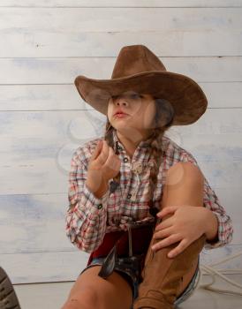 A little girl in a wide-brimmed cowboy hat wearing a traditional dress and high boots with a lasso is eating a straw on a light wooden background.