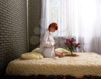 A young red-haired girl sits in the morning on a low bed near the window only in a man's light-colored shirt and drinks coffee.