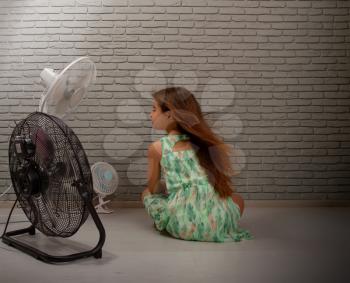 A little girl cools herself from the heat with the help of three different fans at once sitting on the floor of the room