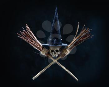 Halloween image Skull in a traditional witch pointed black hat and two crossed brooms on an ominous dark background
