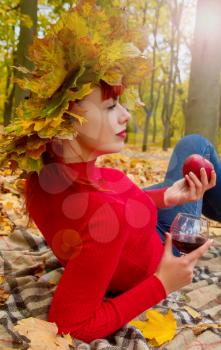 young girl in bright clothes and a wreath of maple leaves lonely and thoughtfully lies in the autumn park with a glass of wine in her hand.