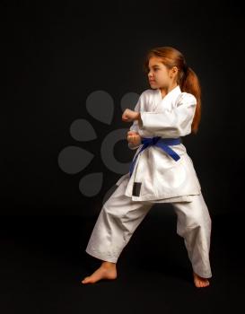 Little girl in a sports kimono and a blue belt performs exercises in kata on a dark background