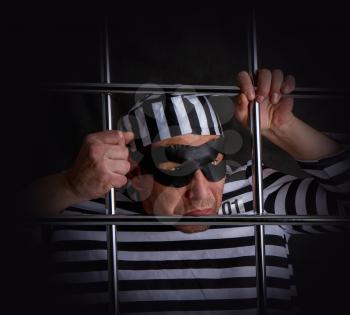 A comic thug in a striped prisoner costume and black mask sits in prison behind a metal bars