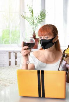 A young girl in a medical mask drinks red wine, communicating with a friend over the Internet on a netbook