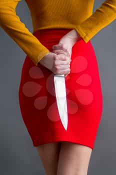 A young girl in a bright skirt and blouse hides a large steel kitchen knife behind a back in her arms on a dark background