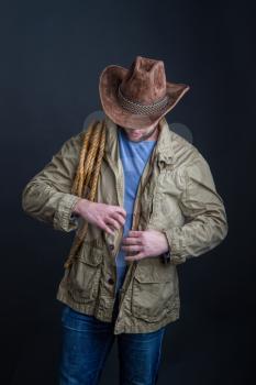 Cowboy in a wide-brimmed hat and jacket with a lasso on his shoulder posing on a dark background
