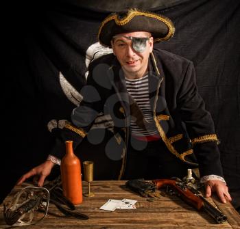an adult pirate in a cocked hat and a striped vest plays cards and a wooden table against the background of the Jolly Roger flag