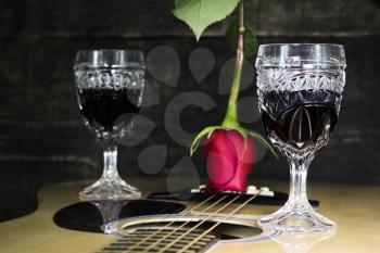 Glass Of Red Wine Resting on Acoustic Guitar With Red Rose and another Glass in The Background 