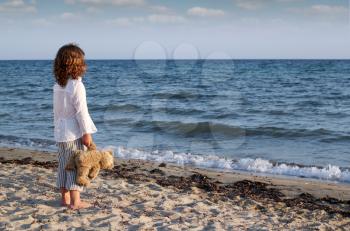 little girl with teddy bear standing on beach and looking at the sea