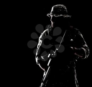 Half length studio contour backlight shot of bearded special forces soldier in uniforms with weapons, portrait on black background.