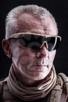 Close up studio shot of special forces white-haired veteran in field uniforms, black background. Protective goggles glasses are on