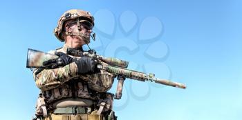 Half length low angle location shot of special forces soldier in field uniforms with weapons, portrait on blue sky background