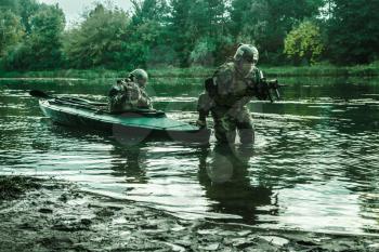Covert landing. Pair of armed operators with painted faces disembarking river coast from military kayak . Diversionary mission under cover of darkness