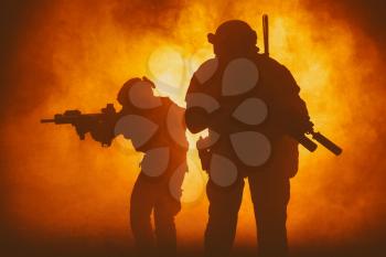 Black silhouettes of pair of soldiers in smoke moving through fire burning in battle operation, sacrificing their lives of our security. Back light