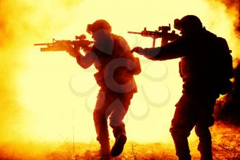 Black silhouettes of pair of soldiers in the smoke fire moving in battle operation. One raising hand to warn another