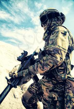Airsoft, strikeball player in camouflage uniform with military tactical ammunition and service carbine replicas climbing on sand dune. Modern war game in sandy area or desert, armed conflict imitation