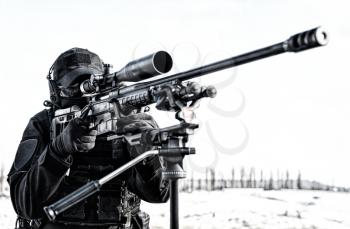 Sniper of police special operations tactical group in black uniforms, ballistic glasses and headphones, aiming with telescopic optical sight on sniper rifle mounted on tripod, over shoulder back view