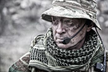 Close-up portrait of brutal commando veteran, experienced army commander or officer with dirty face, wearing camouflage bonnie, shemagh, tactical radio headset with microphone, looking in camera