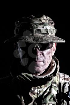 Army veteran, elderly commando fighter, experienced mercenary in camouflage uniform, wearing bonnie and tactical glasses, looking in camera, studio, low key shoulder portrait on black background