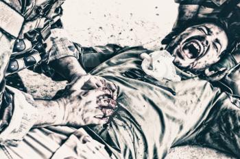 Wounded soldier, commando fighter lying on floor, screaming in pain while comrade pressing with hands on wound in his stomach, trying stop bleeding and save life. Tactical casualty care on battlefield