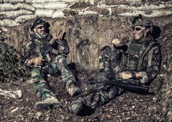 Navy SEALs soldiers, special operations forces fighters in camouflage uniform, body armor and helmet, armed assault rifles, sitting in trench, resting after fight, talking and joking with comrades