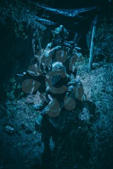 Two army soldiers, Navy SEALs team shooters wearing combat uniform, body armor and helmet, aiming service rifles, covering each other while moving in trench. Infantrymen on battlefield at night