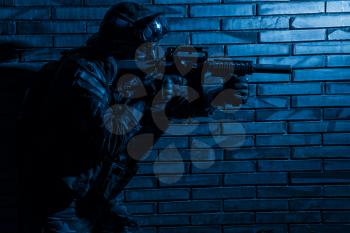 Army soldier, counter terrorist squad fighter in camouflage uniform, load carrier and goggles on helmet, sneaking in darkness along brick wall, aiming service rifle with silencer during indoor fight