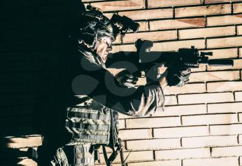 Army soldier, counter terrorist squad fighter in camouflage uniform, load carrier and goggles on helmet, sneaking in darkness along brick wall, aiming service rifle with silencer during indoor fight