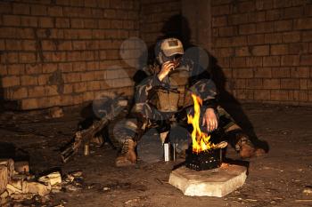 Special forces soldier after the fight sitting by the fire in ruined building smoking cigarette