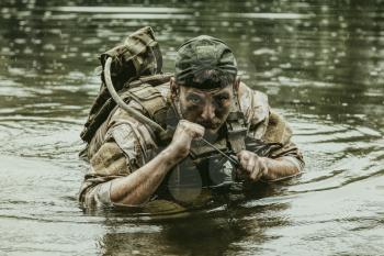 Private military contractor PMC in baseball cap during river raid in the jungle waist deep in the water and mud