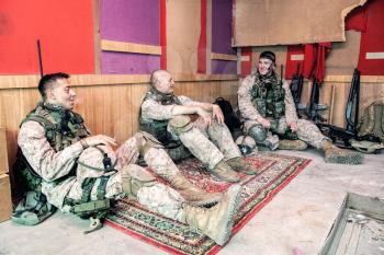 United States marines in camouflage uniform and ammunition sitting on floor at combat outpost or temporary base on mission, talking in relaxing atmosphere, resting after hard day on military service