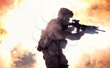 Silhouette of modern infantry soldier, elite army fighter in tactical ammunition and helmet, standing with assault service rifle in hands on background of fiery explode. Burning fire of war conflict
