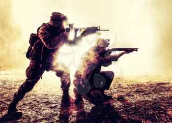 Silhouettes of two army soldiers, U.S. marines team in action, surrounded fire and smoke, shooting with assault rifle and machine gun, attacking enemy with suppressive gunfire during offensive mission