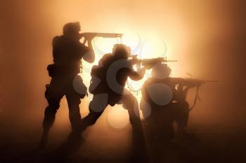 Silhouettes of three army soldiers, U.S. marines team in action, surrounded fire and smoke, shooting with assault rifle and machine gun, attacking enemy with suppressive gunfire during offensive mission
