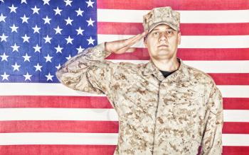 Portrait of U.S. army soldier in camouflage uniform and cap saluting on background of flag of United States of America, looking at camera. Military hand salute to display respect for national flag
