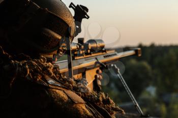 Special forces sniper with large-caliber rifle seeking and killing enemy. Back view sunset sky background