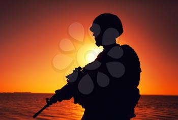 Silhouette of army infantry soldier, special forces rifleman armed with service rifle, patrolling coastline at sunset. Coast guard fighter in combat helmet and tactical ammunition standing on seashore