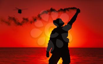 Silhouette of army special operations forces soldier, commando fighter signaling, marking landing spot or evacuation area for helicopter pilot with smoke flair while standing on shore during sunset
