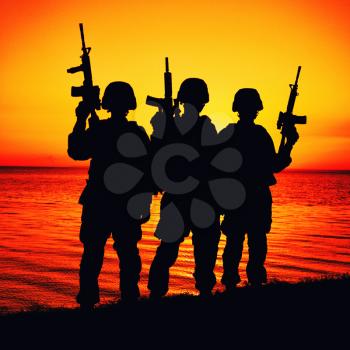 Silhouette of army special operations forces soldiers team, group of Marines or coast guard fighters crew in ammunition and combat helmets standing on seashore at sunset with raised assault rifles