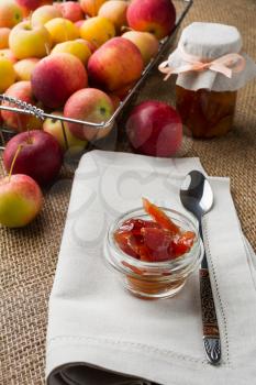 Glass jar of apple confiture, linen napkin and apples in metal wire basket on a burlap covered table, vertical, vertical. Selective focus. The toning
