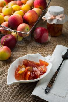Homemade apple slices jam on white saucer, spoon, jar of marmalade, linen napkin and apples in metal wire basket on a burlap covered table, vertical. Selective focus. The toning